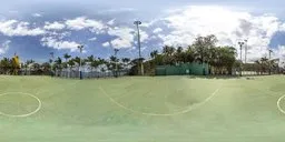 360-degree HDR panorama of a sunlit football field, surrounded by fencing and palms, with clear skies ideal for scene lighting.