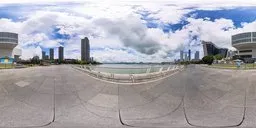 Overcast sky over a coastal plaza with modern buildings, yachts, statues, and lush greenery.