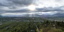 Aerial HDR panorama featuring sunbeams piercing through clouds above a verdant valley with river and mountain backdrop.